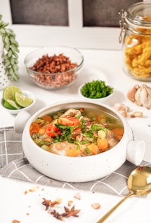 A ceramic bowl filled with a colorful soup, including ingredients like carrots, tomatoes, and herbs. The soup is garnished with fresh greens. Surrounding the bowl are various ingredients such as lime wedges, chopped greens in a small dish, garlic cloves, uncooked rice in a glass bowl, and a jar of crispy snacks. A gold spoon is placed nearby on a checkered napkin, and star anise and dried onion flakes are scattered around.