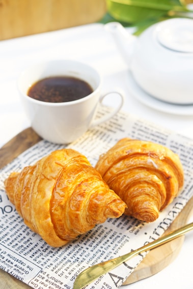 A warm, golden croissant resting on a rustic wooden table beside a steaming cup of specialty coffee.