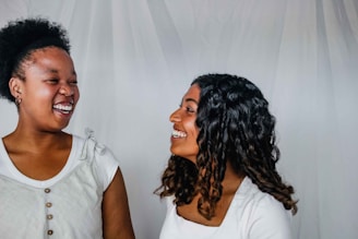 Two people are facing each other and smiling, creating a joyful and friendly interaction against a plain backdrop. Both are wearing light-colored tops, and their hair is styled in different ways, with one having curly hair and the other with short, natural hair.