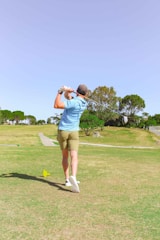 A person is playing golf on a sunny day, wearing a light blue shirt, khaki shorts, and a cap. They are in the middle of swinging a golf club on a well-maintained golf course. The surrounding area features green grass, a few trees, and an open sky.