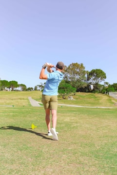 A person is playing golf on a sunny day, wearing a light blue shirt, khaki shorts, and a cap. They are in the middle of swinging a golf club on a well-maintained golf course. The surrounding area features green grass, a few trees, and an open sky.