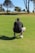 A person in athletic attire crouches on a golf green, focusing on a small golf ball nearby. The setting includes a flagstick positioned on the green and a backdrop of large trees under a clear sky.