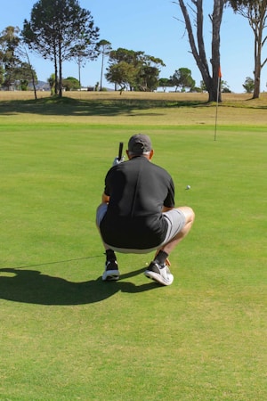 A person in athletic attire crouches on a golf green, focusing on a small golf ball nearby. The setting includes a flagstick positioned on the green and a backdrop of large trees under a clear sky.
