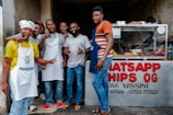 Seven men stand in front of a food stand, some wearing aprons and one dressed as a chef. The group appears cheerful and is posing with thumbs up. A glass display case showing various food items is visible, and a sign reads 'WHATSAPP CHIPS OG.'