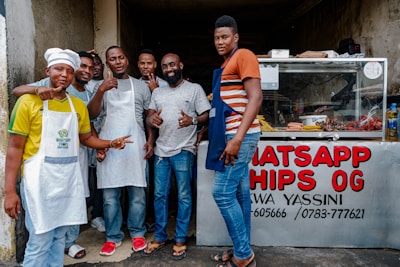 Seven men stand in front of a food stand, some wearing aprons and one dressed as a chef. The group appears cheerful and is posing with thumbs up. A glass display case showing various food items is visible, and a sign reads 'WHATSAPP CHIPS OG.'