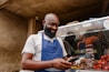 A man with a beard and a bald head is wearing a blue apron over a light gray shirt. He is looking at his smartphone while smiling. Behind him, there is a food stall with various grilled meats. Another two people can be seen in the reflections on the glass of the food stall.
