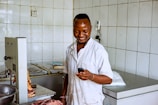 A person in a white lab coat is standing in a room with tiled walls, holding a smartphone and smiling. The room contains equipment and surfaces that suggest a work environment, possibly related to food preparation or processing. Pieces of raw meat are visible on a metal surface.