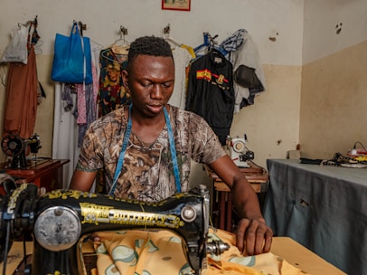 A man is sitting at a table operating a sewing machine. He is wearing a camouflage shirt and has a blue measuring tape around his neck. Behind him, there are several garments hanging on a rack, along with various sewing materials on the tables and walls of the room. The scene appears to be a sewing or tailoring workshop with a variety of clothing fabrics and sewing equipment.