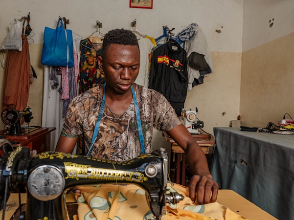 A man is sitting at a table operating a sewing machine. He is wearing a camouflage shirt and has a blue measuring tape around his neck. Behind him, there are several garments hanging on a rack, along with various sewing materials on the tables and walls of the room. The scene appears to be a sewing or tailoring workshop with a variety of clothing fabrics and sewing equipment.