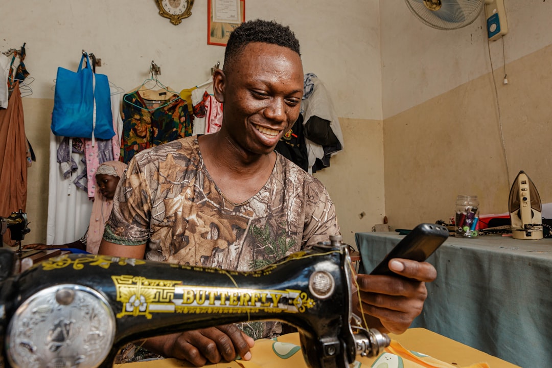 a man smiles as he uses a sewing machine, African tailor using his smartphone happy smiling