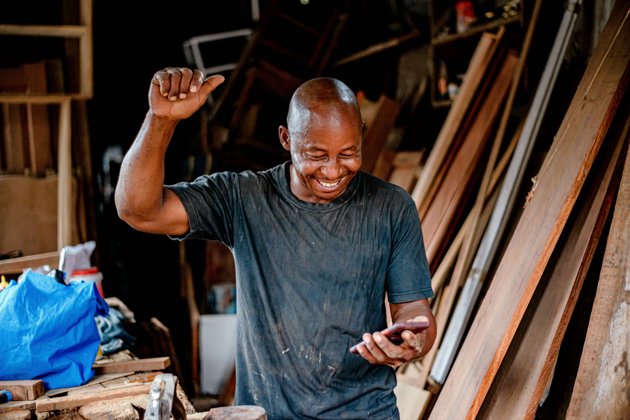 Carpenter laughs and punches the air while reading his phone in a timber-filled workshop.