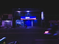 A night scene featuring a building with illuminated signage displaying the logo and name 'BRI' in bright blue and red colors. The street in front appears empty and a few ATM machines are visible through the glass frontage. A motorcycle is blurred in motion across the foreground.