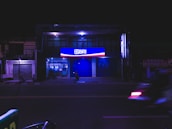 A night scene featuring a building with illuminated signage displaying the logo and name 'BRI' in bright blue and red colors. The street in front appears empty and a few ATM machines are visible through the glass frontage. A motorcycle is blurred in motion across the foreground.