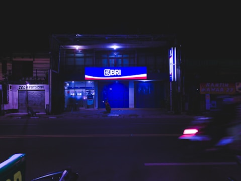 A night scene featuring a building with illuminated signage displaying the logo and name 'BRI' in bright blue and red colors. The street in front appears empty and a few ATM machines are visible through the glass frontage. A motorcycle is blurred in motion across the foreground.