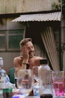A man is sitting at a table with a hand on his face, smiling and appearing amused. Surrounding him are various bottles and glasses, suggesting a social setting. In the background, there is a corrugated metal wall and a partially open curtain.