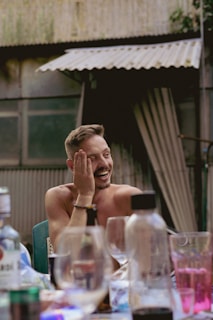 A man is sitting at a table with a hand on his face, smiling and appearing amused. Surrounding him are various bottles and glasses, suggesting a social setting. In the background, there is a corrugated metal wall and a partially open curtain.