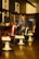 Three classic barber chairs with red and white upholstery are lined up in a barber shop. The interior features wooden paneling and mirrored stations with various hair products. Black-and-white portraits are mounted on the wall, adding a vintage atmosphere. The overall setting exudes a warm and nostalgic ambience.