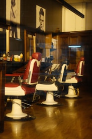 Three classic barber chairs with red and white upholstery are lined up in a barber shop. The interior features wooden paneling and mirrored stations with various hair products. Black-and-white portraits are mounted on the wall, adding a vintage atmosphere. The overall setting exudes a warm and nostalgic ambience.