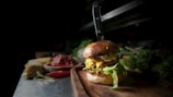A gourmet burger with a knife inserted into it is displayed on a wooden board surrounded by fresh lettuce and various ingredients. The background includes elements such as raw meat, corn, and chili peppers, suggesting a kitchen or dining setting. The lighting highlights the textures and layers of the burger, including cheese and leafy greens.