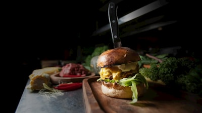 A gourmet burger with a knife inserted into it is displayed on a wooden board surrounded by fresh lettuce and various ingredients. The background includes elements such as raw meat, corn, and chili peppers, suggesting a kitchen or dining setting. The lighting highlights the textures and layers of the burger, including cheese and leafy greens.
