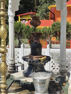 A traditional Hindu Shiva Lingam setup is featured, surrounded by ornate architectural details. The Lingam is black with a metal pot hanging above it, presumably for ceremonial water or milk pouring. A white, intricately designed fence and lush green trees are visible in the background, with colorful buildings providing a vivid contrast. A metal bucket filled with liquid is placed in front of the Lingam.
