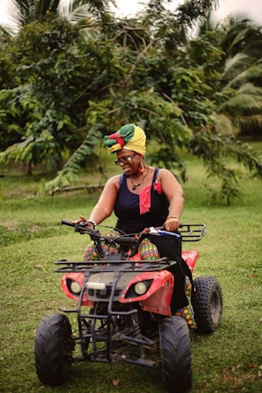 A person wearing a multicolored head wrap and a black top is riding a red ATV across a grassy area. The individual is smiling and appears to be enjoying the ride. The background is filled with green foliage and palm trees.
