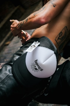 A shirtless person with tattoos is holding a cigarette. A black and white cap with the word 'bones' and a skull design rests on their knee. Leather pants and chain detail are visible.