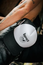 A close-up of a person's tattooed arm, wearing a white and black cap with a design that includes a skull and a spider web with the word 'bones' on it. The person appears to be wearing black leather pants.
