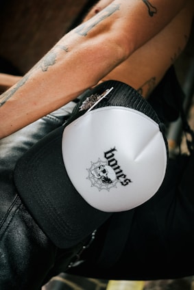 A close-up of a person's tattooed arm, wearing a white and black cap with a design that includes a skull and a spider web with the word 'bones' on it. The person appears to be wearing black leather pants.