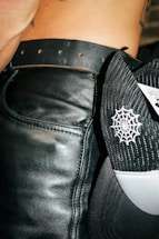 A close-up view of a person wearing a black leather belt around their waist. Next to the belt is a black and white mesh cap with a spider web pattern embroidered on it. Part of the person's skin and tattoo is visible.