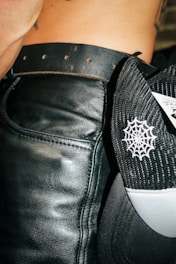 A close-up view of a person wearing a black leather belt around their waist. Next to the belt is a black and white mesh cap with a spider web pattern embroidered on it. Part of the person's skin and tattoo is visible.