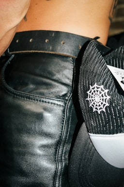 A close-up view of a person wearing a black leather belt around their waist. Next to the belt is a black and white mesh cap with a spider web pattern embroidered on it. Part of the person's skin and tattoo is visible.