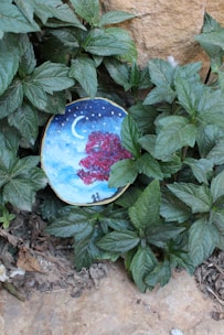 A small circular artwork depicting a night scene with a crescent moon and stars, featuring a prominent red tree and two silhouettes of people standing under it. The artwork is placed among lush green leaves and is partially encircled by them, with a stone backdrop visible.