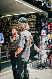 Two individuals stand outside a shop filled with various hats and sunglasses. One person with curly hair is wearing a graphic t-shirt, leather pants, and a cap with a skull design. The other person has short hair, wears a patterned button-up shirt and black pants. The shopfront is vibrant with colors from the hats and sunglasses on display.