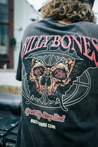 A person with curly hair is wearing a t-shirt featuring a striking design with bold text and a large graphic of a moth. The words 'BILLYBONES,' 'Speak Up Stay Bad,' and 'BILLYBONES CLUB' are prominently displayed in various stylized fonts. The surrounding environment includes an industrial or urban setting, with blurred architectural elements in the background.