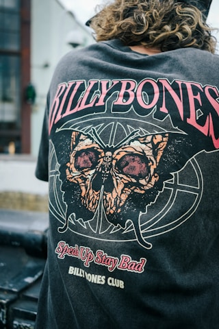 A person with curly hair is wearing a t-shirt featuring a striking design with bold text and a large graphic of a moth. The words 'BILLYBONES,' 'Speak Up Stay Bad,' and 'BILLYBONES CLUB' are prominently displayed in various stylized fonts. The surrounding environment includes an industrial or urban setting, with blurred architectural elements in the background.