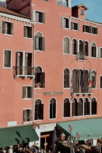 A large building with a pink facade features multiple windows with dark shutters and small balconies. The name 'Hotel Rialto' is prominently displayed on the building. Several flags are visible hanging from one of the balconies. The lower part of the building has a green awning, and some people are gathered around the entrance.