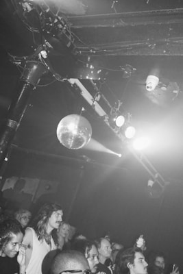A crowded scene at a nightclub featuring a diverse group of people watching something off-camera. The ceiling is adorned with a disco ball and stage lights, creating a lively and energetic atmosphere.