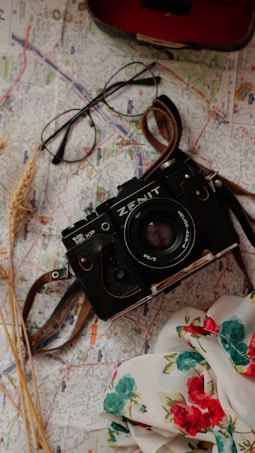 A vintage black camera with 'Zenit' branding rests on a detailed street map. Next to the camera, a pair of black-framed eyeglasses sits slightly askew. A floral scarf with red and teal flowers occupies the bottom right corner, while a few stalks of wheat are placed to the left. The overall arrangement suggests a travel or exploration theme.