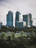 Several skyscrapers under construction dominate the skyline, with cranes visible at the top of each building. In the foreground, a park with lush greenery and a few people walking. The scene suggests a blend of urban development and nature.