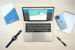 A laptop computer displaying a website on its screen is placed on a light wooden surface. Nearby are a blue smartphone, a smartwatch with a dark blue band, a blue folder, a white notepad, and a black pen.