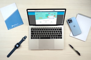 A laptop computer displaying a website on its screen is placed on a light wooden surface. Nearby are a blue smartphone, a smartwatch with a dark blue band, a blue folder, a white notepad, and a black pen.
