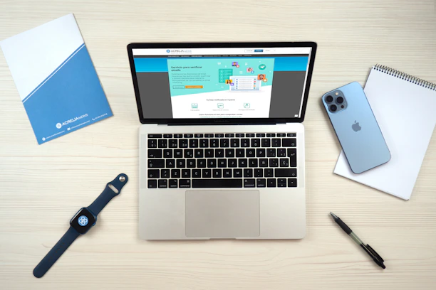 A laptop computer displaying a website on its screen is placed on a light wooden surface. Nearby are a blue smartphone, a smartwatch with a dark blue band, a blue folder, a white notepad, and a black pen.