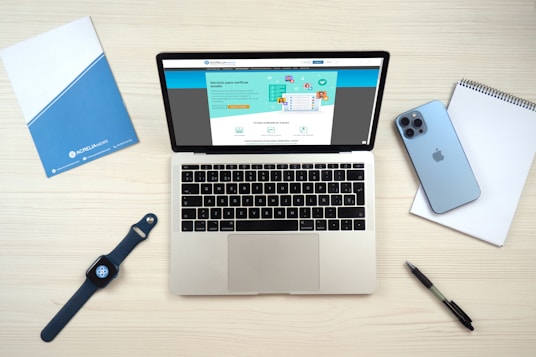 A laptop computer displaying a website on its screen is placed on a light wooden surface. Nearby are a blue smartphone, a smartwatch with a dark blue band, a blue folder, a white notepad, and a black pen.