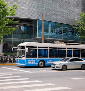 A blue and white bus and a silver car are stopped at a crosswalk in an urban setting. The background features a modern building with glass windows displaying an illuminated outline of a car charging. Several motorcycles are parked along the sidewalk, and there are green leafy trees on either side of the scene.