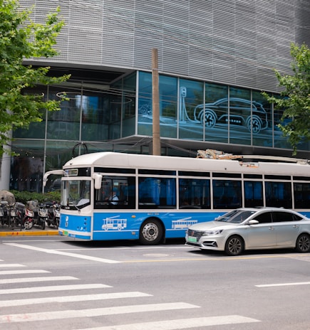 A blue and white bus and a silver car are stopped at a crosswalk in an urban setting. The background features a modern building with glass windows displaying an illuminated outline of a car charging. Several motorcycles are parked along the sidewalk, and there are green leafy trees on either side of the scene.