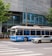 A blue and white bus and a silver car are stopped at a crosswalk in an urban setting. The background features a modern building with glass windows displaying an illuminated outline of a car charging. Several motorcycles are parked along the sidewalk, and there are green leafy trees on either side of the scene.