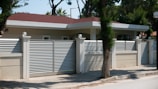 A modern house with a flat roof is surrounded by a high beige fence with metal slats and decorative spherical lights on top of the posts. It is located on a paved street with trees and greenery in the background.