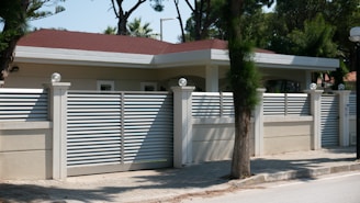 A modern house with a flat roof is surrounded by a high beige fence with metal slats and decorative spherical lights on top of the posts. It is located on a paved street with trees and greenery in the background.