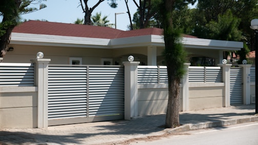 A modern house with a flat roof is surrounded by a high beige fence with metal slats and decorative spherical lights on top of the posts. It is located on a paved street with trees and greenery in the background.